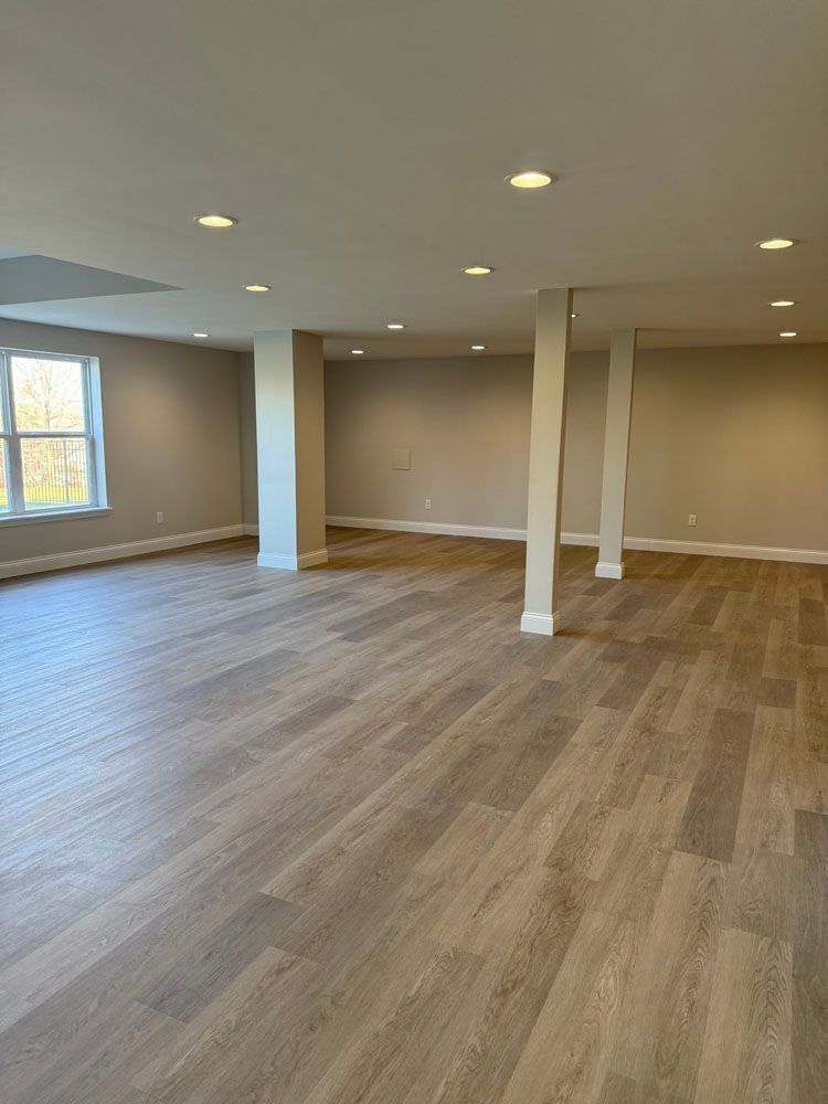 Empty basement room with light wood-look flooring, neutral walls, recessed lighting, support columns, and a window.