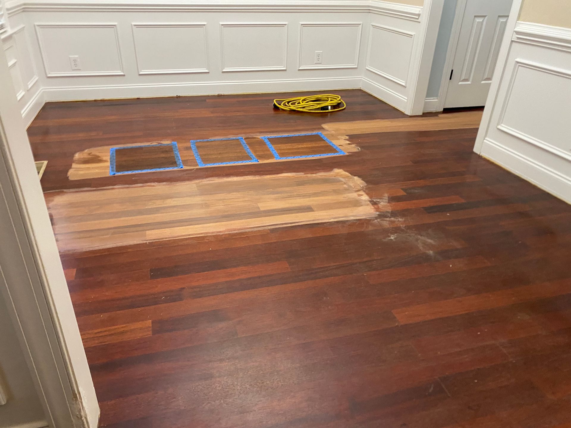 Wooden floor with taped blue and beige repair sections in a white-paneled room; a tape measure sits nearby