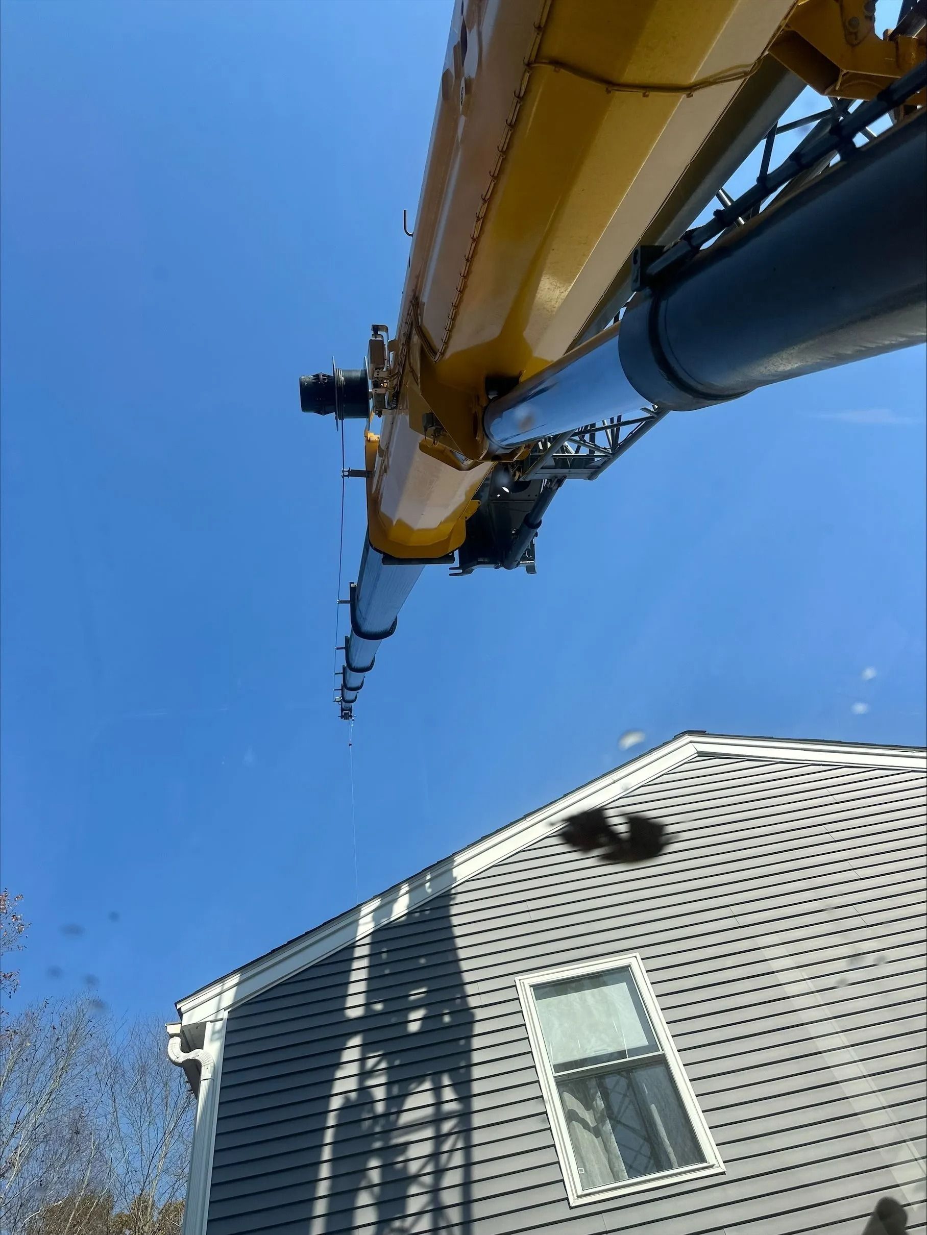 Yellow crane boom extended over a gray house against a blue sky.