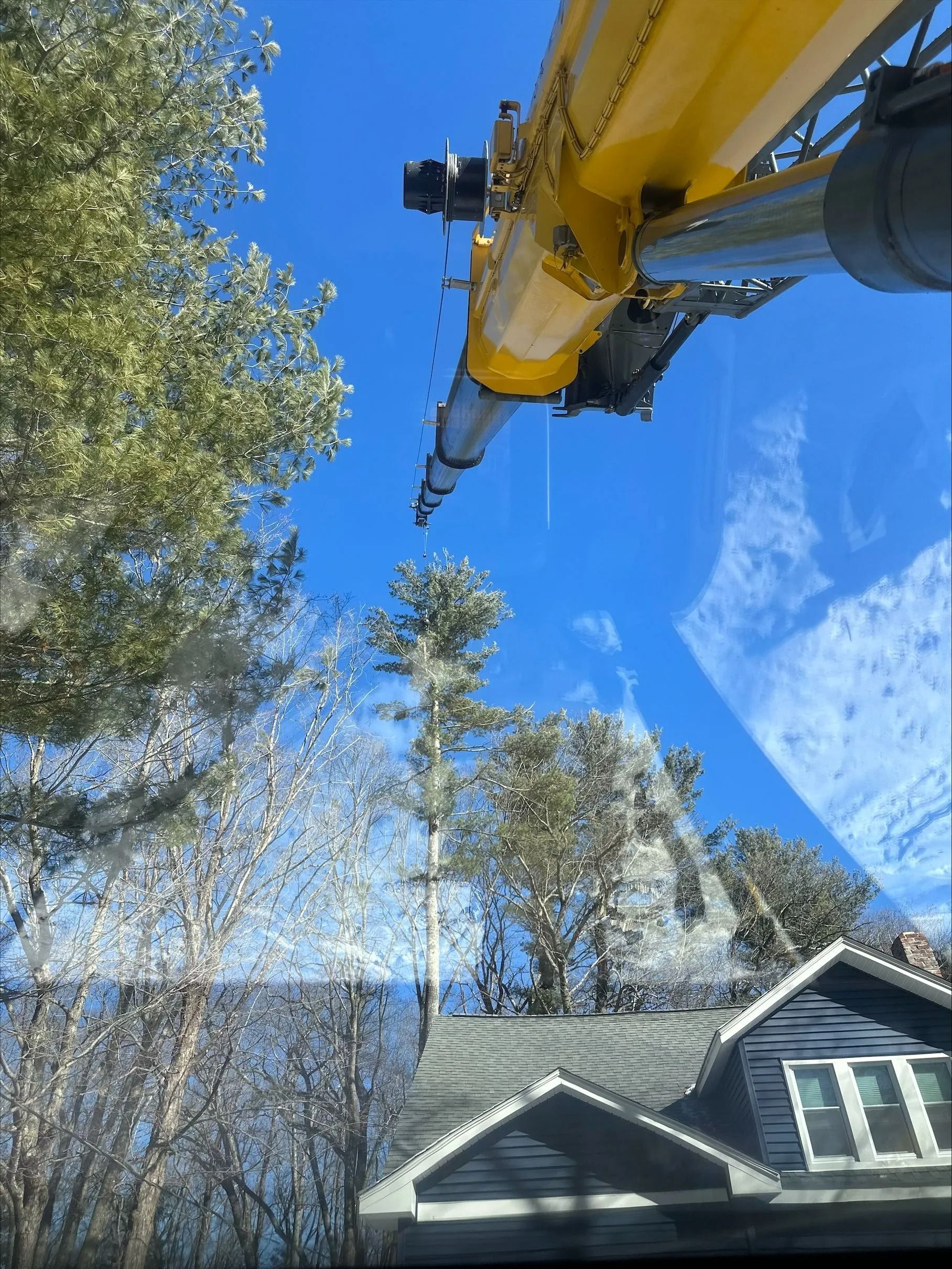 Yellow crane boom over a house and trees under a blue sky.