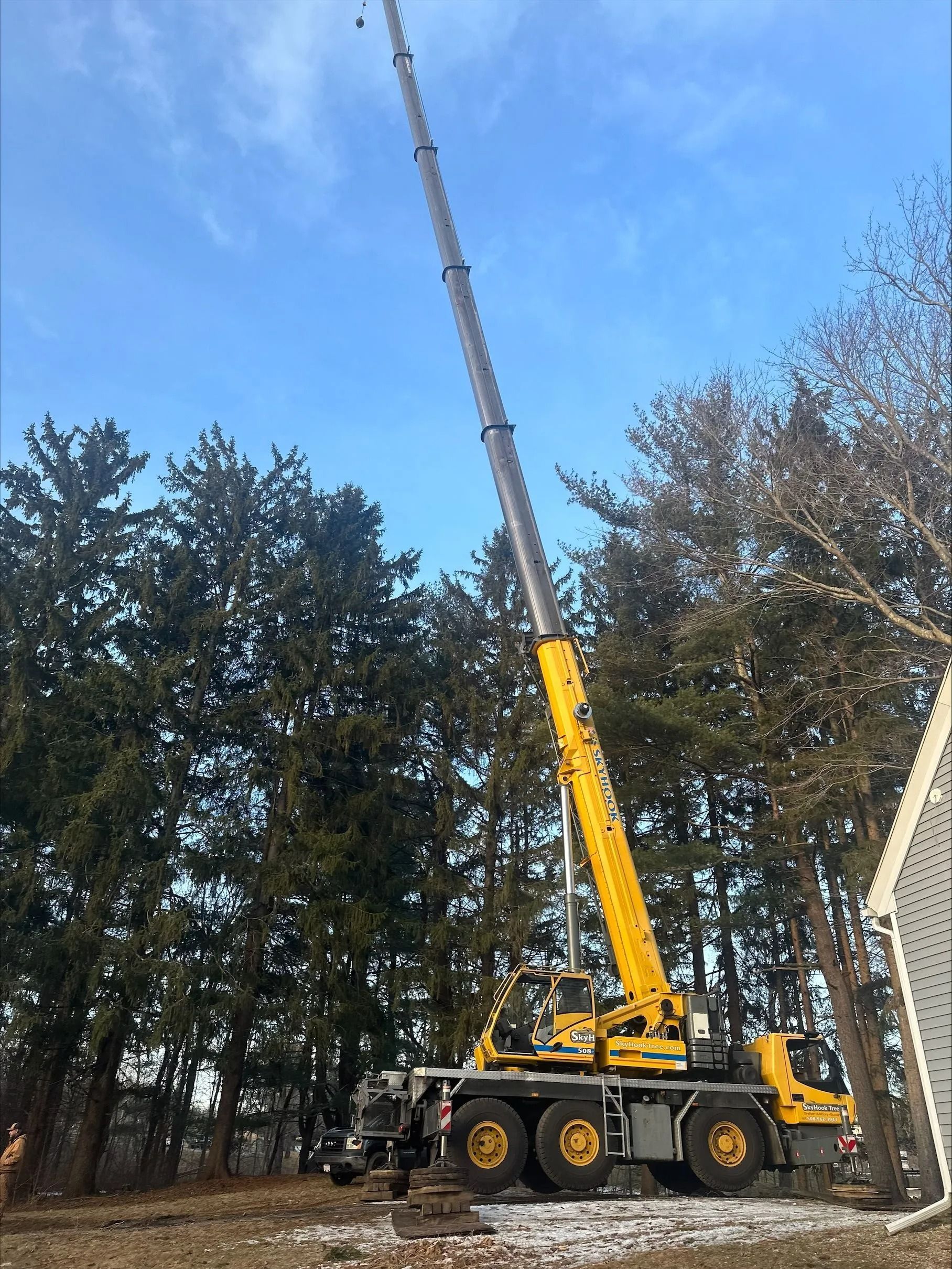 Yellow mobile crane with extended boom in a field near trees, under a blue sky.