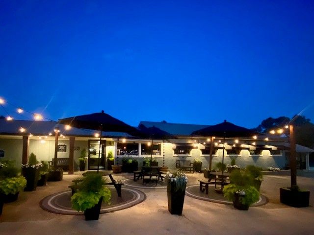 Outdoor patio at dusk with tables, umbrellas, and warm string lights under a blue sky