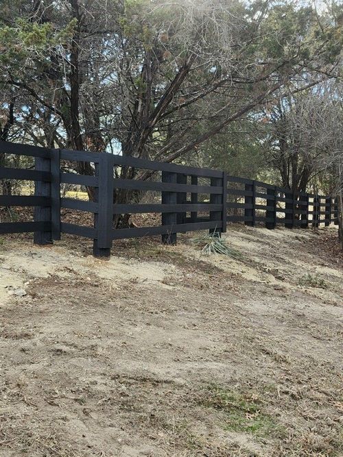 Black wooden fence along a dirt path beside trees and dry grass.