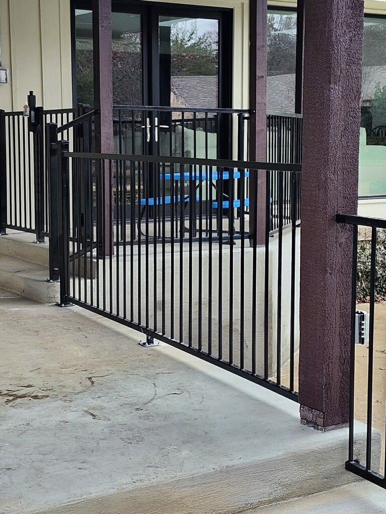 Black metal gate beside a concrete walkway outside a building, with blue patio chairs in the background