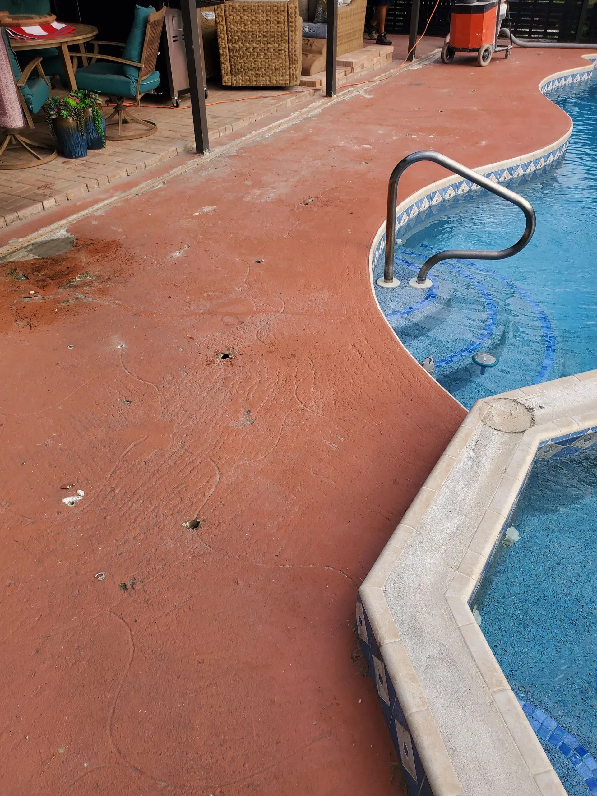 Red concrete pool deck surrounds a blue pool. Handrail on side. Outdoor setting.