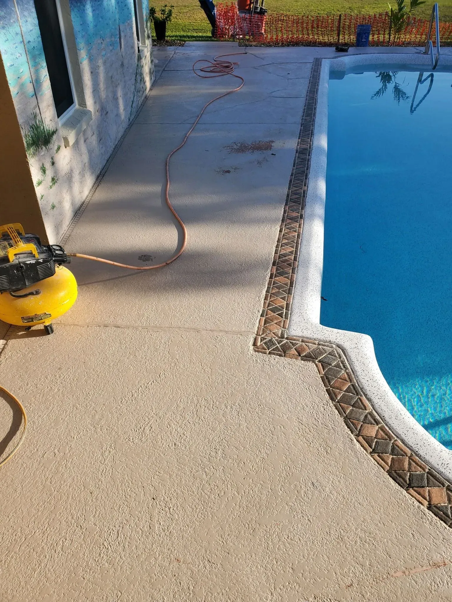 Concrete pool deck next to a blue pool. A hose and air compressor are on the deck.