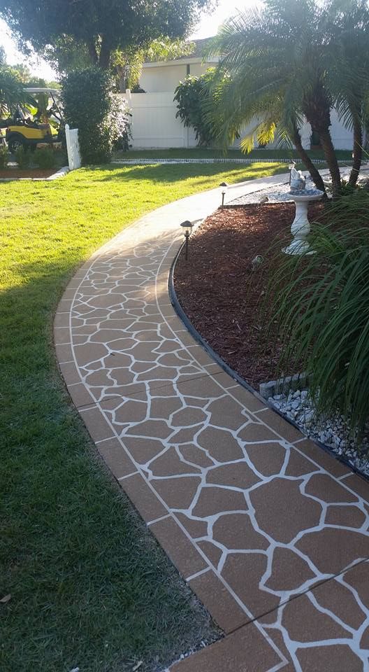 Stone-patterned walkway through a grassy yard, bordered by dark brown mulch and greenery, leading to a white building.