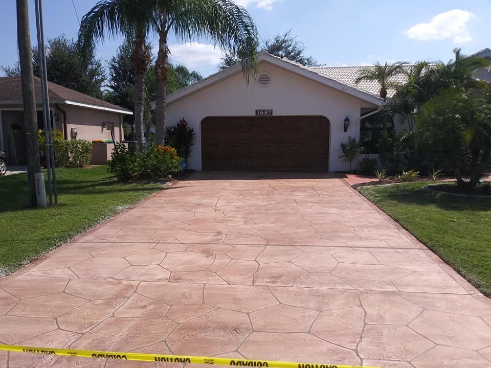 A driveway with a textured pattern leads to a beige house with a brown garage door, on a sunny day.