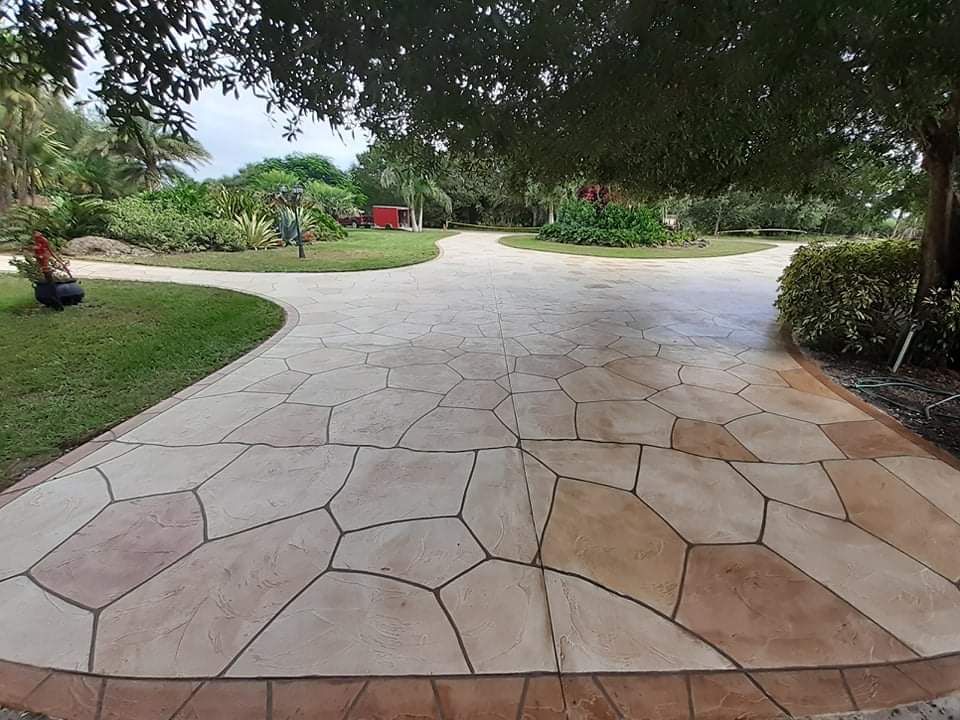 Stone-patterned driveway splitting into two paths, leading to greenery. Trees, grass, and a red structure are in the distance.