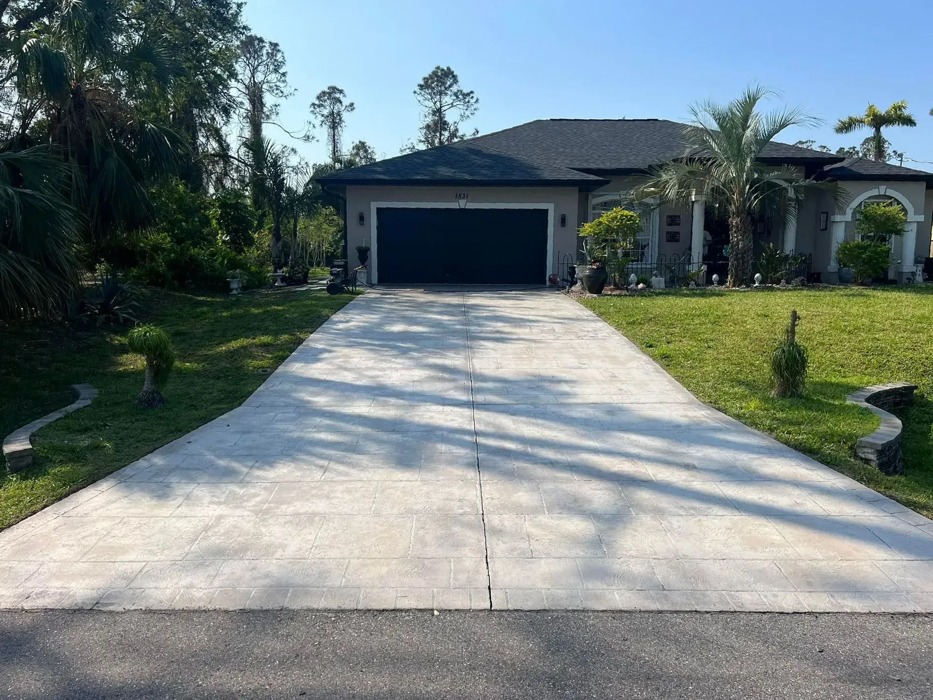 Concrete driveway leading to a gray house with black garage door. Green lawn surrounds.
