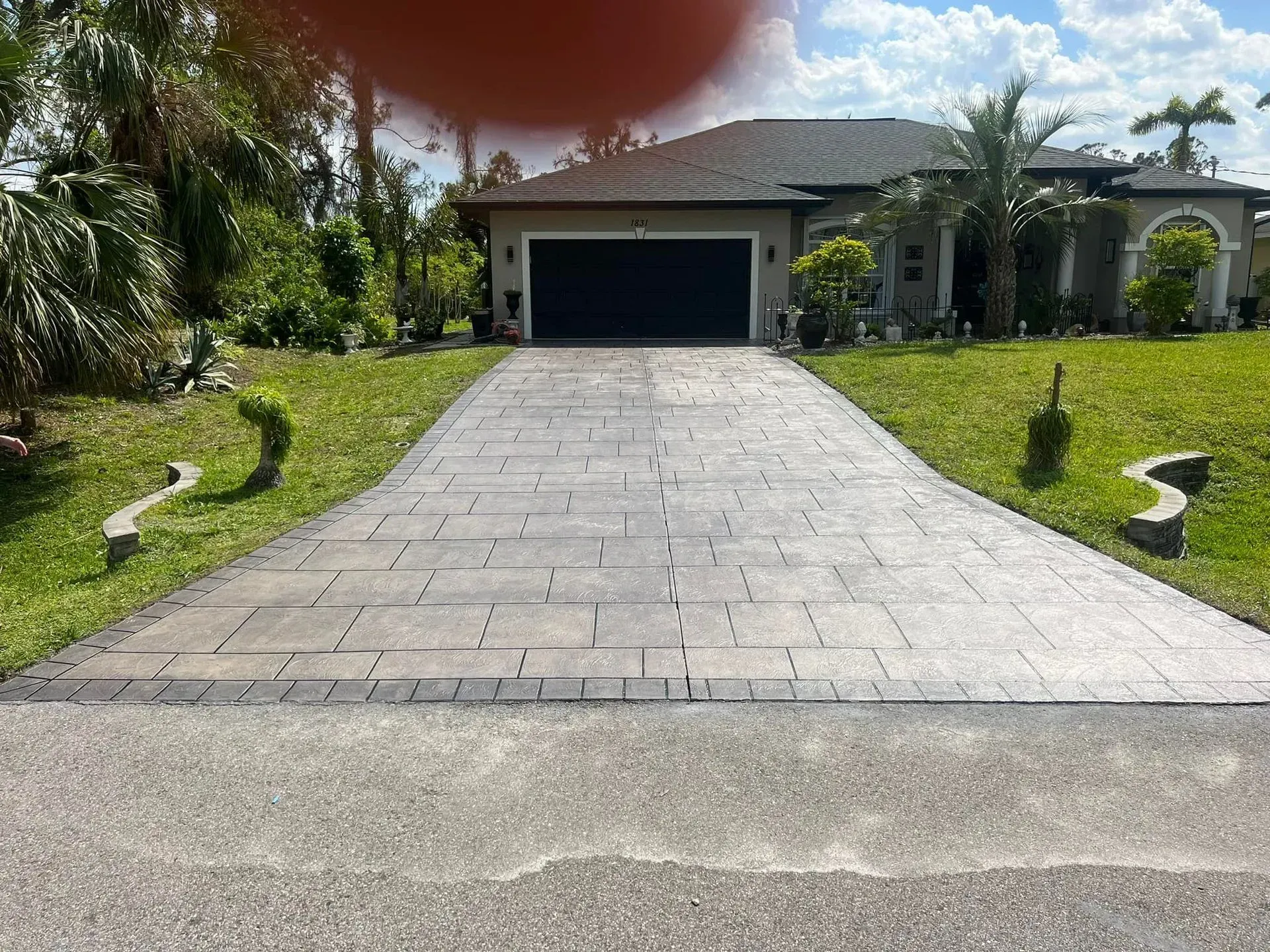 Driveway leading to a house with a black garage door. Green lawn, blue sky, and trees.