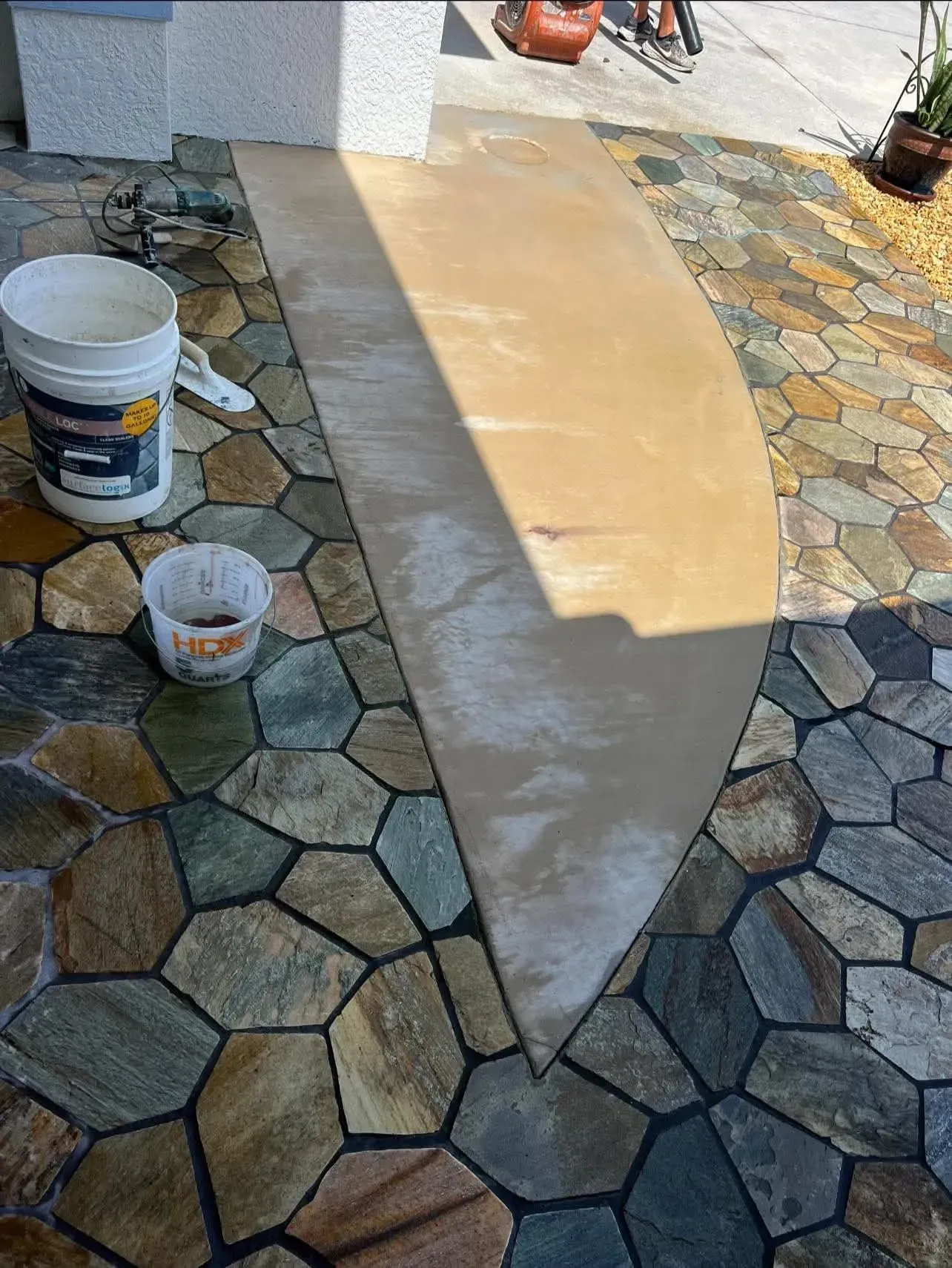 A partially paved entrance with a stone pattern border. A bucket, tools, and fresh concrete are visible.