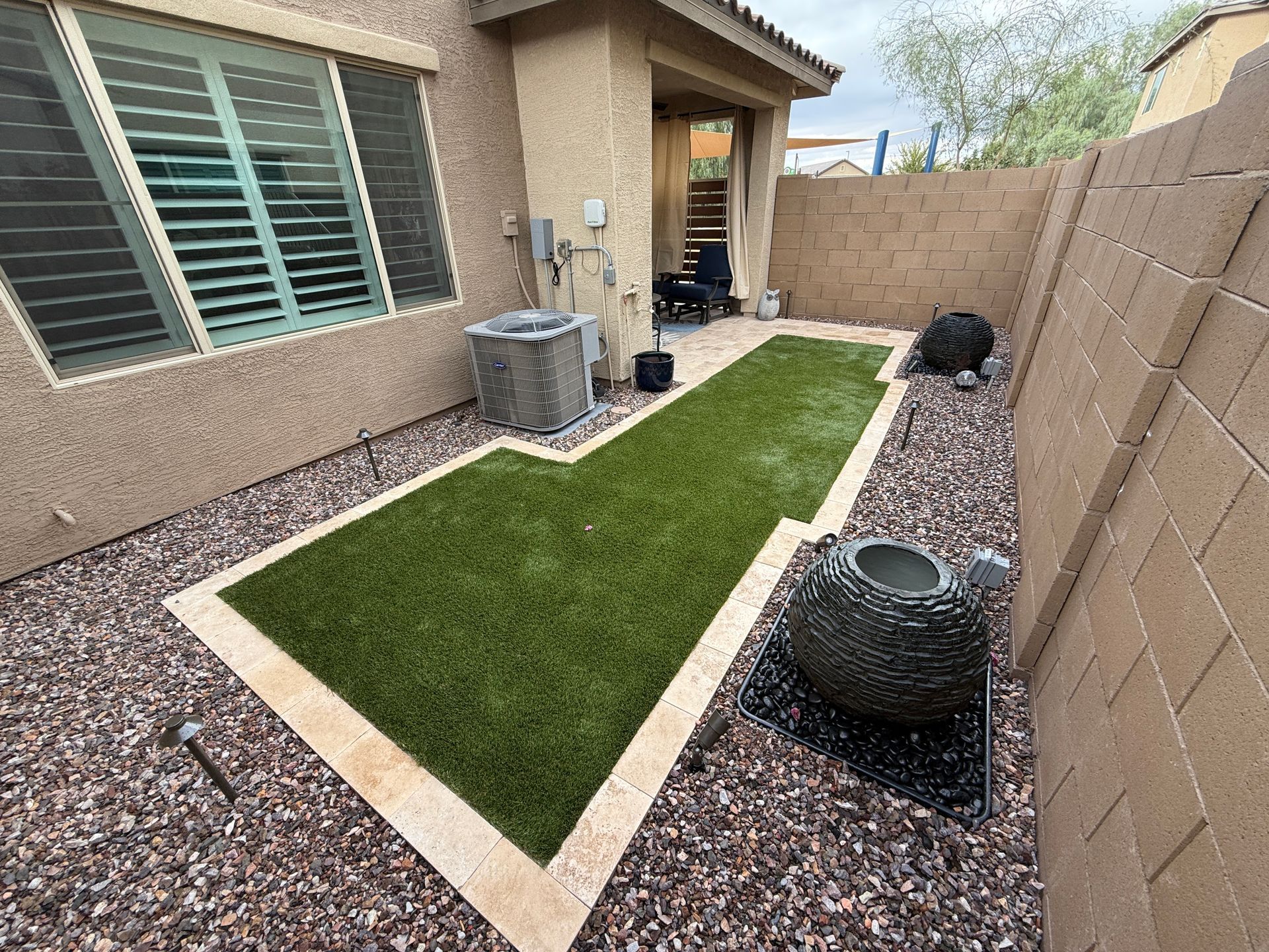 Backyard with green artificial turf, rock borders, beige walls, and water feature.