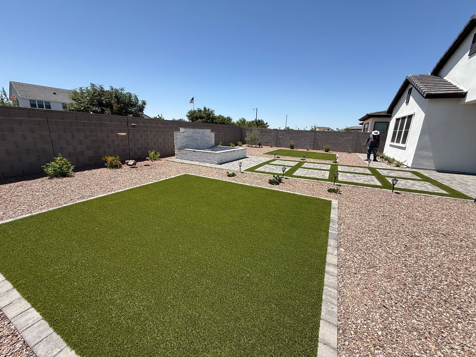 Backyard with artificial turf, gravel, and concrete stepping stones under a blue sky; person near the house.