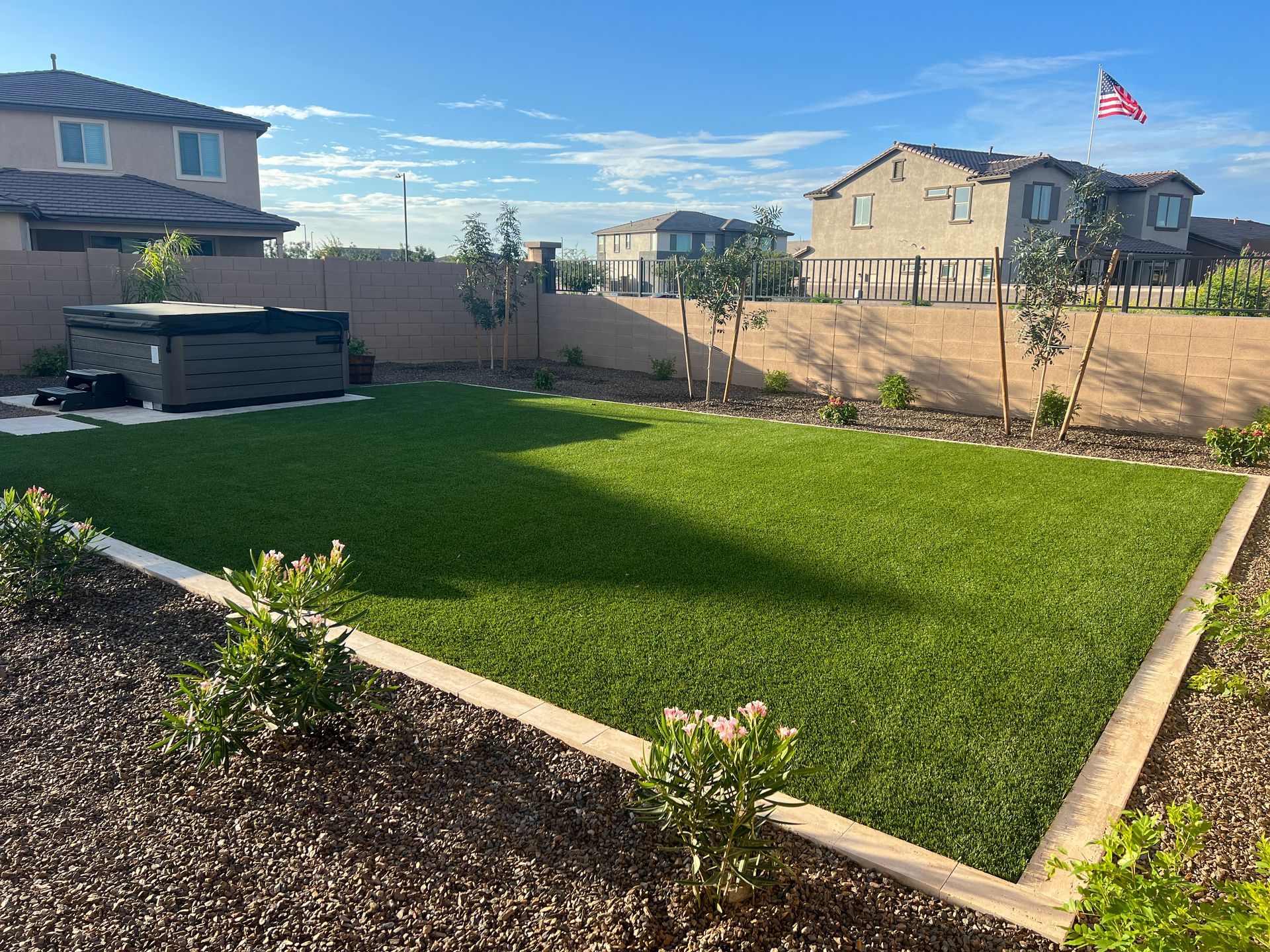 Backyard with green turf, hot tub, small trees, and surrounding homes under a blue sky.