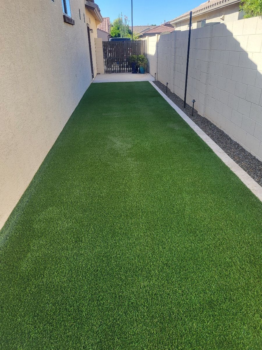 Narrow backyard with green artificial turf between two light-colored walls, leading to a wooden gate.