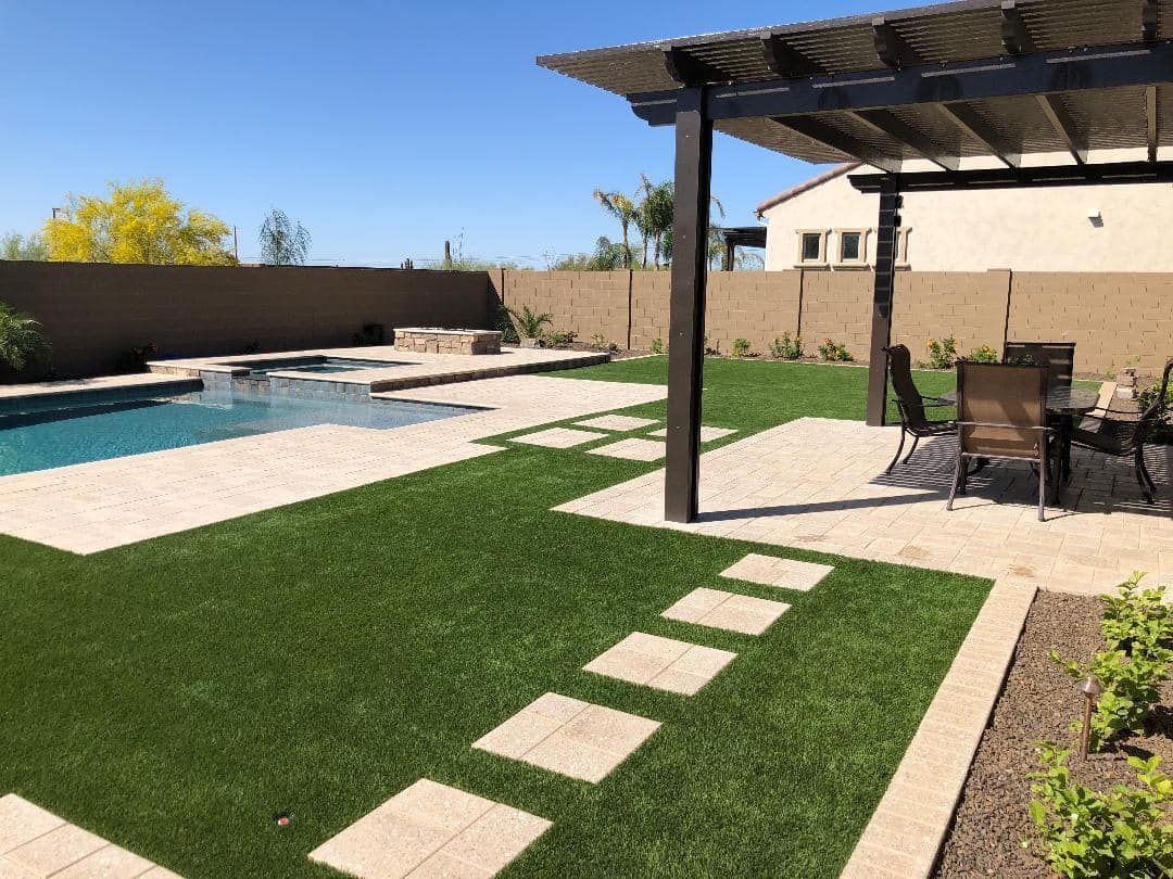 Patio with brown pergola and stone features, next to a beige house and green lawn under a blue sky.