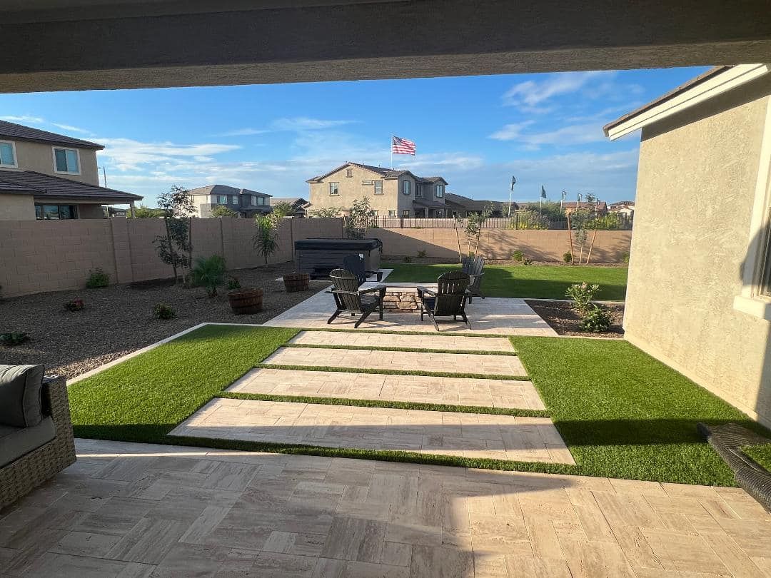 Patio with a fire pit and seating, bordered by grass, with houses and blue sky in the background.