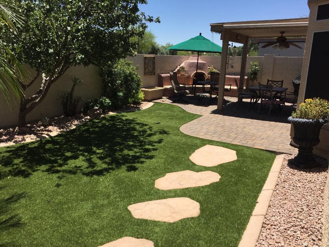 Backyard with green artificial turf, stone path, patio seating under pergola, and brick oven.