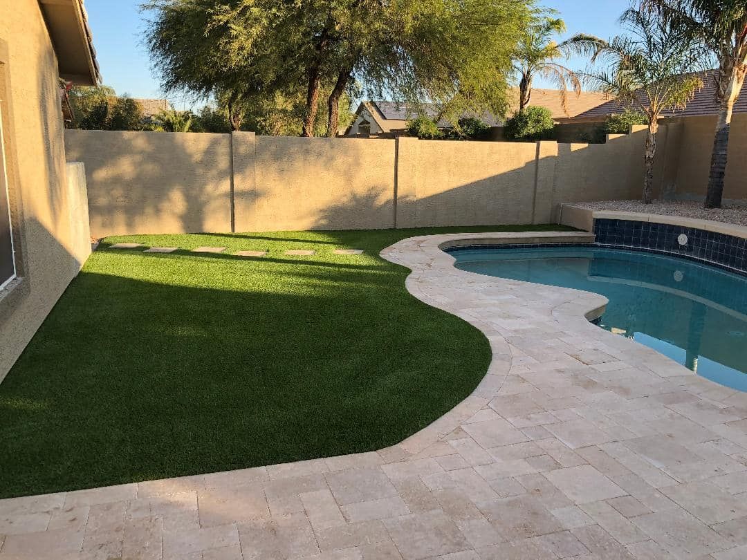Backyard with a swimming pool, green grass, and beige stone patio. A tall wall is in the background.