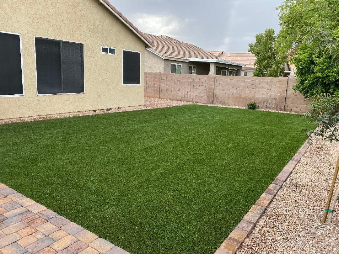 Backyard with green turf surrounded by a brick border and a tan stucco house.