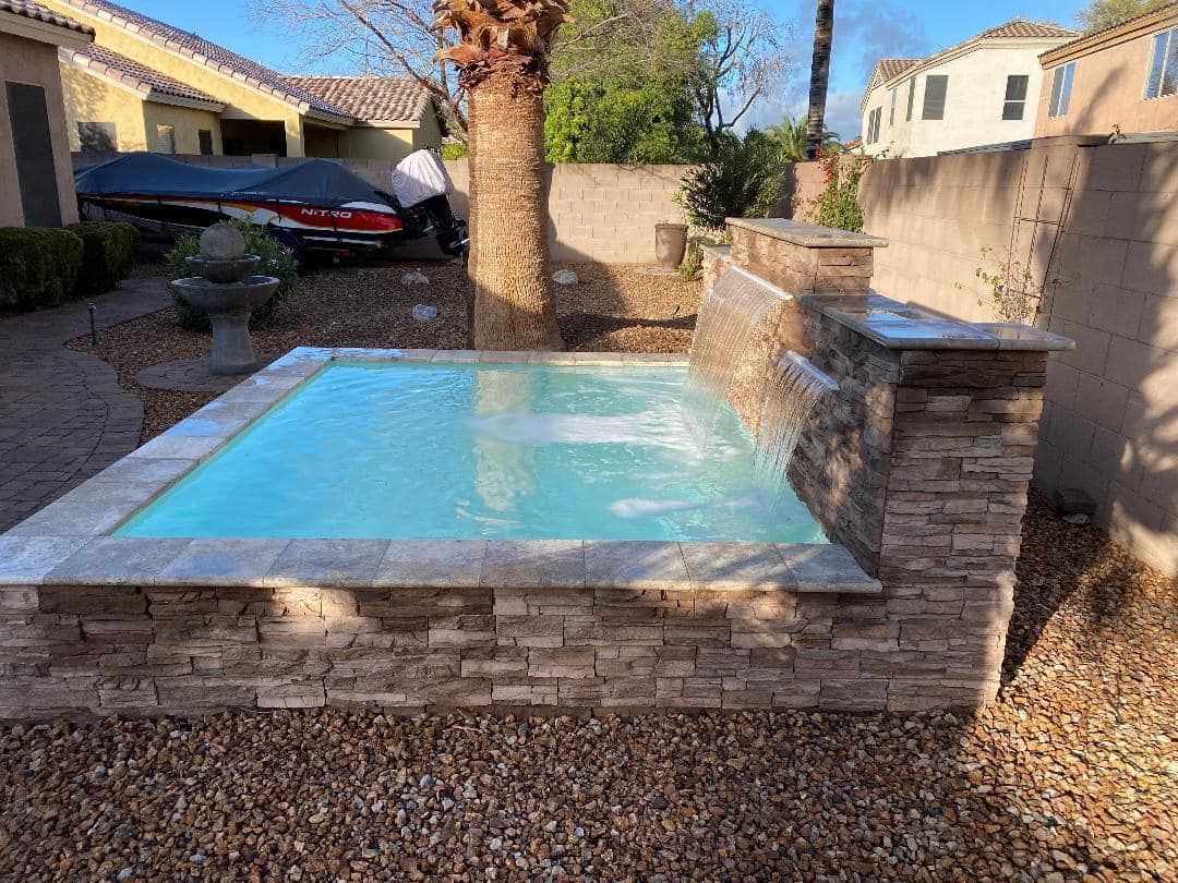 Small rectangular pool with waterfall feature, surrounded by stone and gravel, in a backyard setting.