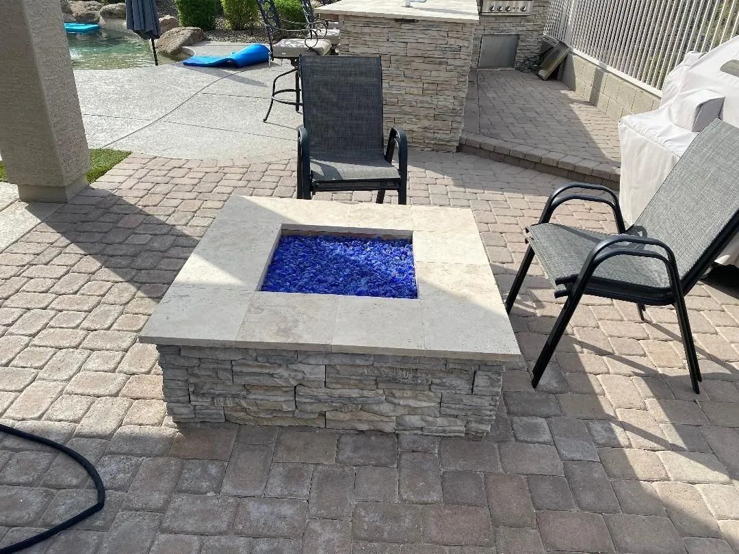 A square stone fire pit with blue glass and two chairs on a patio.