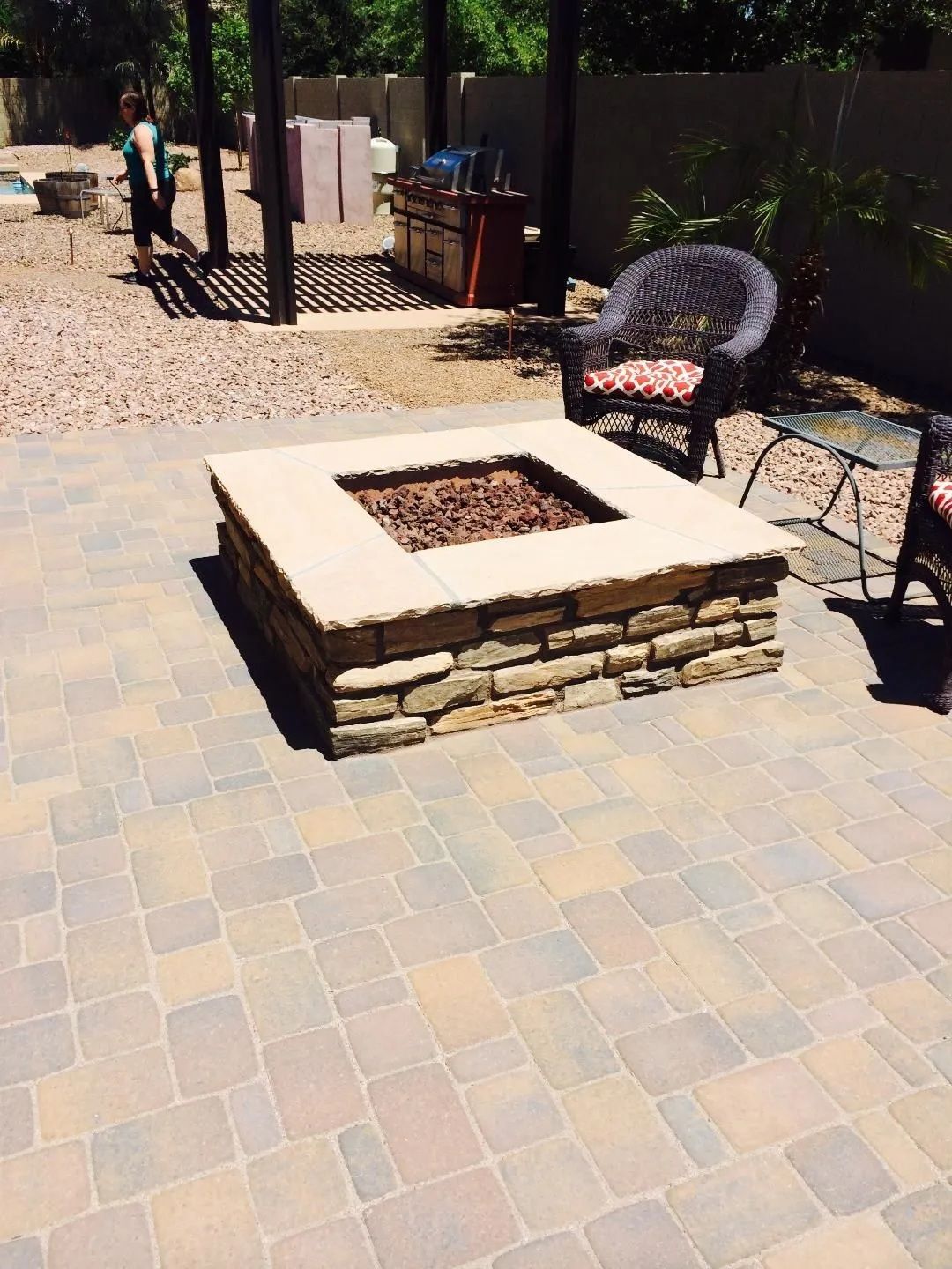 Stone fire pit on a patio. Person walks near a shaded seating area in a backyard.