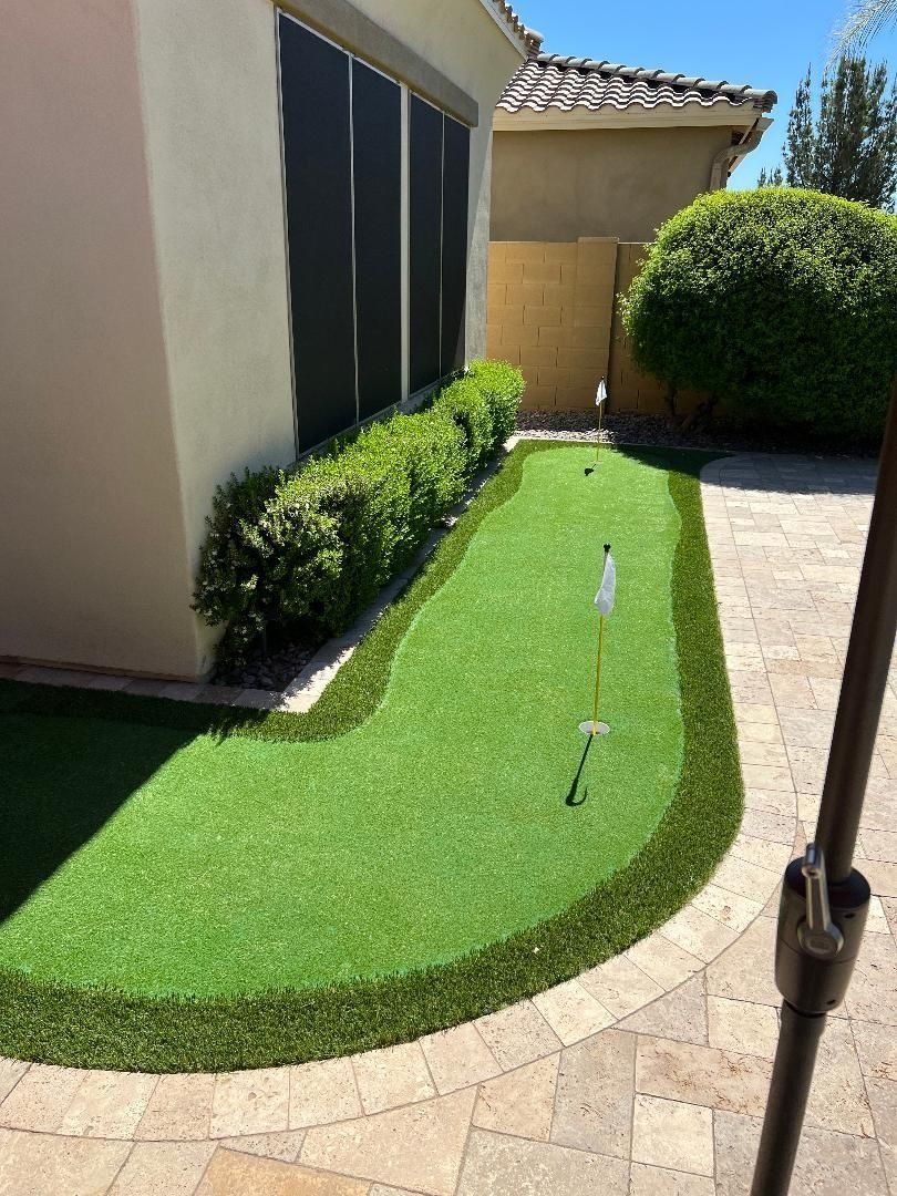 Artificial turf putting green in a backyard, with a flag and bordering green grass. Beige patio.