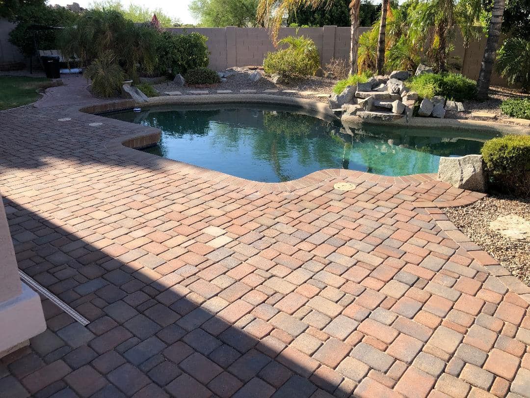 Brick patio surrounds a turquoise pool in a backyard with landscaping and a rock waterfall.
