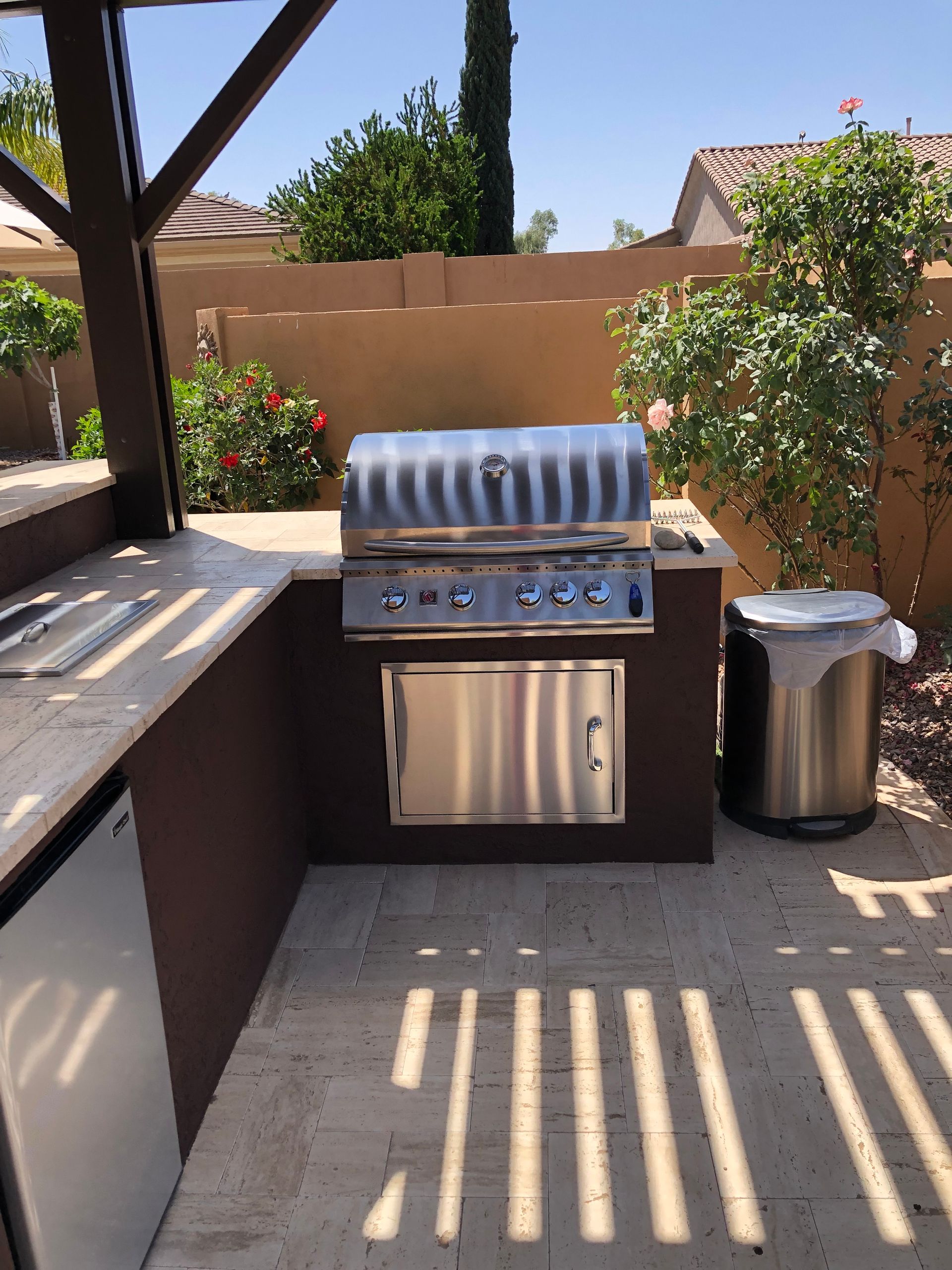 Outdoor built-in grill with stainless steel features, side counter, and trash can. Set within a patio with shade.