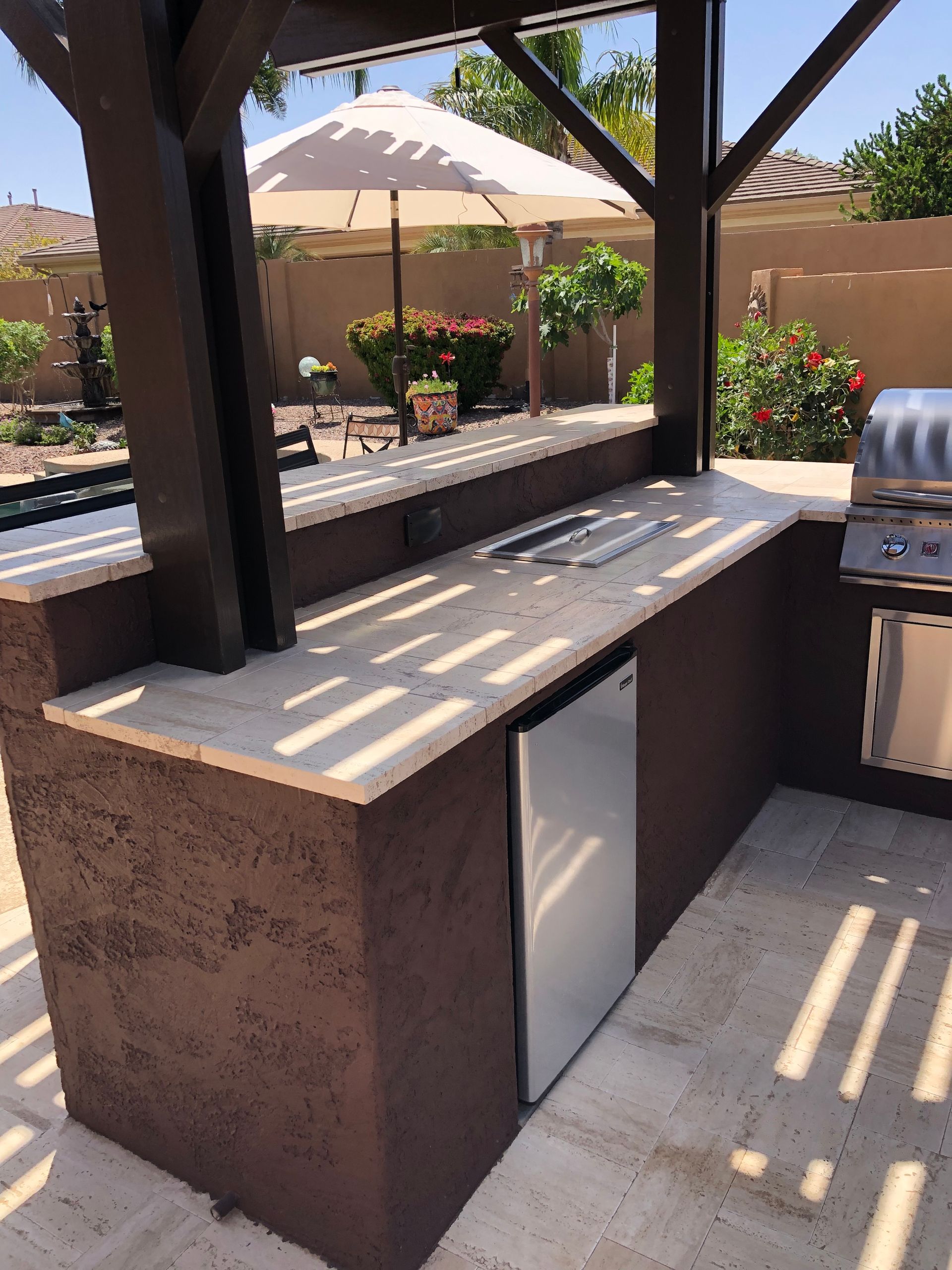 Outdoor kitchen with grill, refrigerator, and countertop under a pergola.