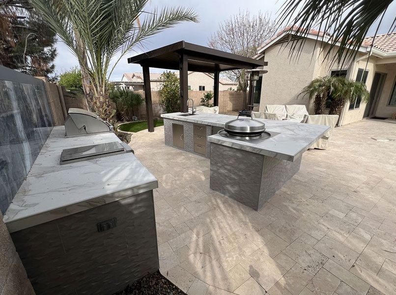Outdoor kitchen with gray and white countertops and a wooden pergola in a backyard.