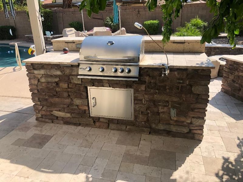 Outdoor stone-clad grill island with stainless steel grill and storage door, on a patio next to a pool.