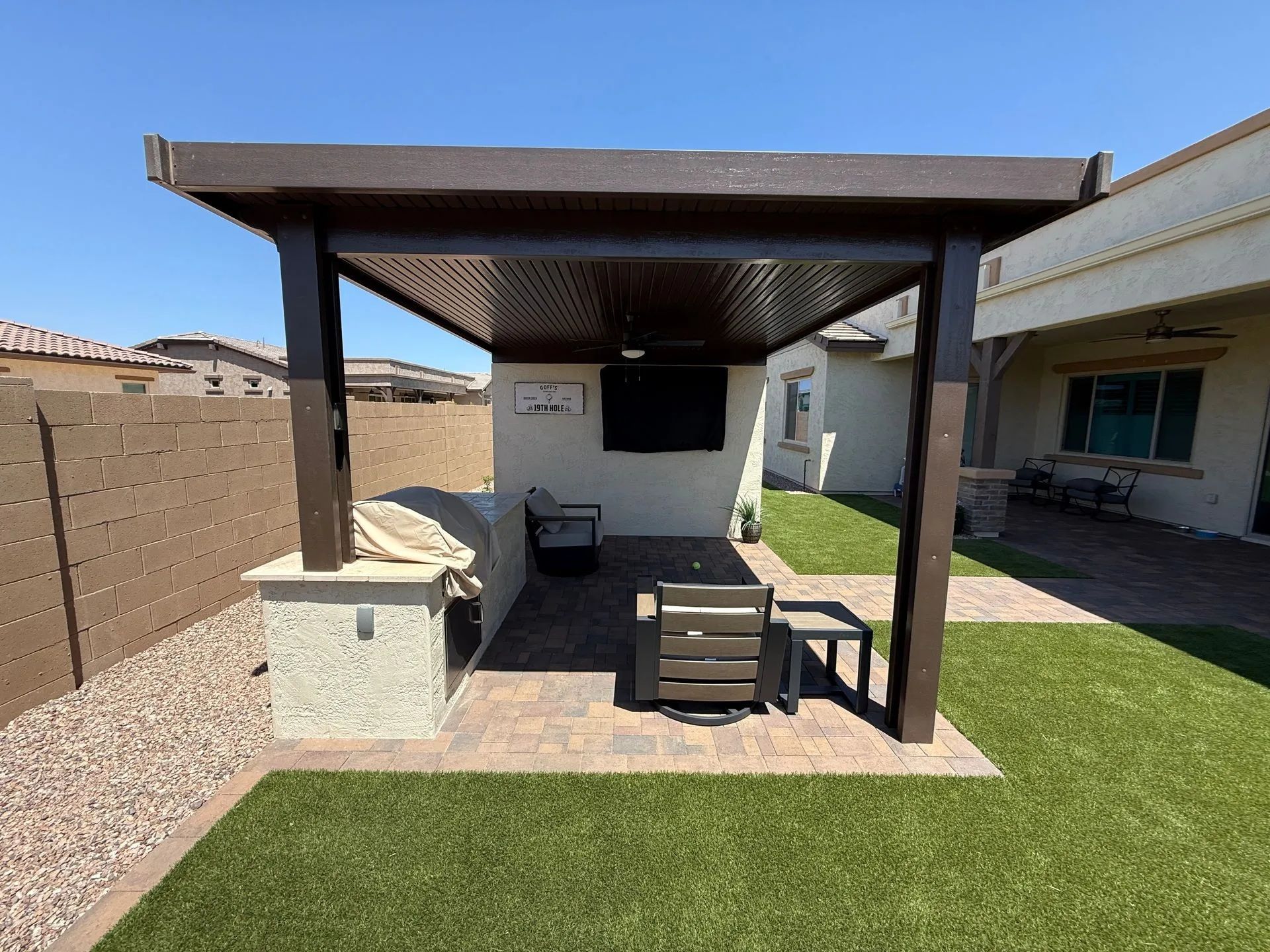 Backyard patio with brown pergola, outdoor TV, grill, and seating on brick pavers.