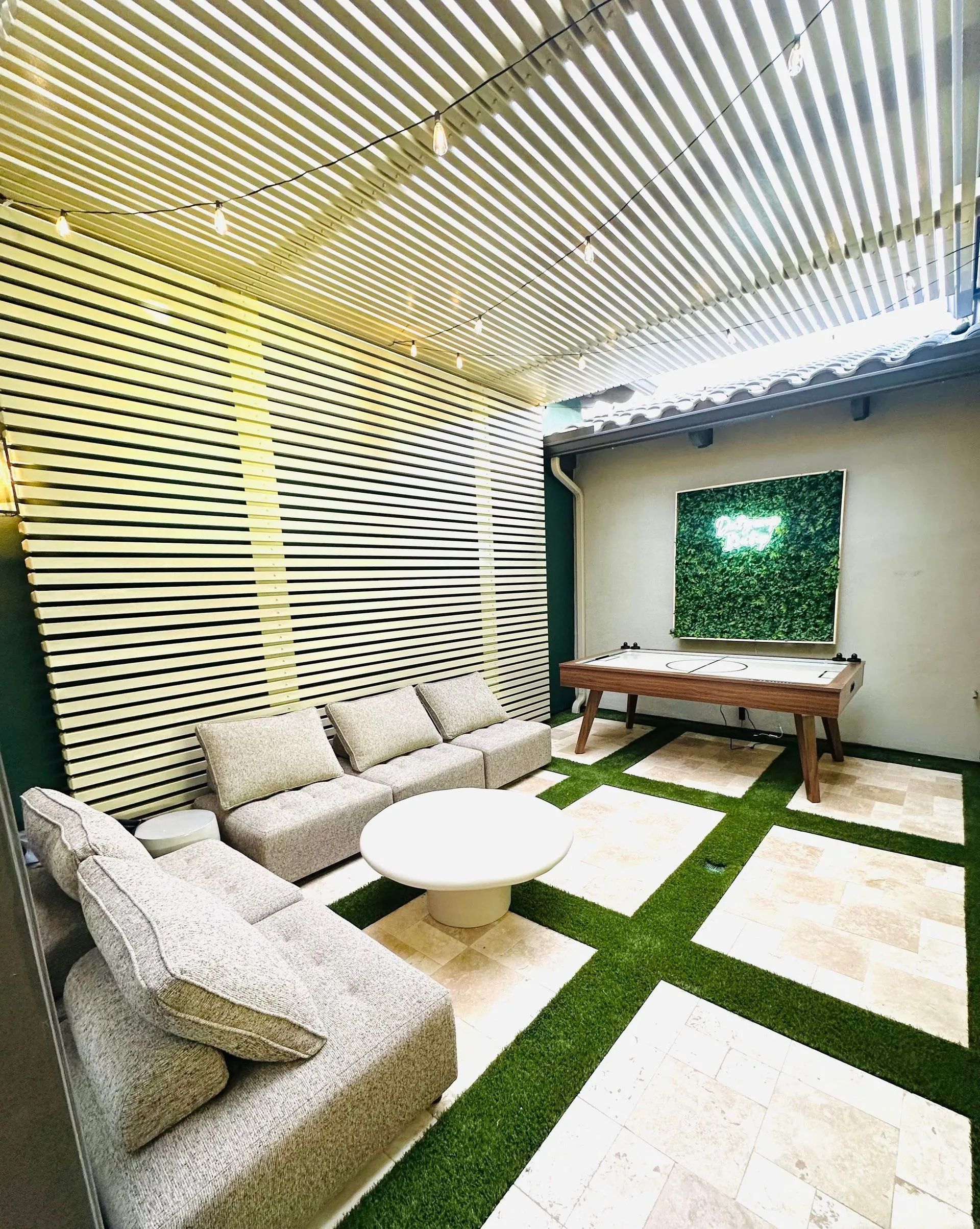 Outdoor lounge area with sofa, coffee table, and pool table. Artificial turf and decorative wall.