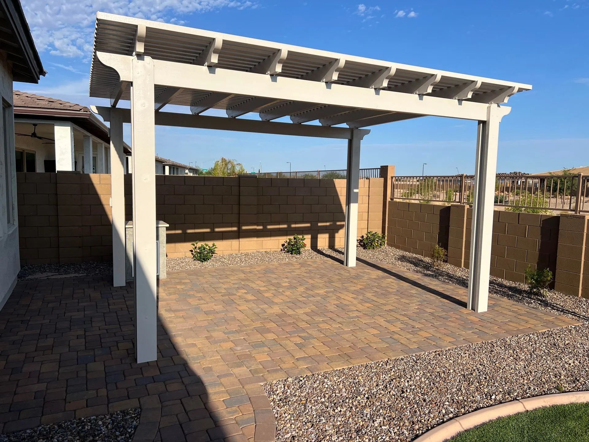 White pergola over a brick patio in a backyard setting with a stone wall and blue sky.