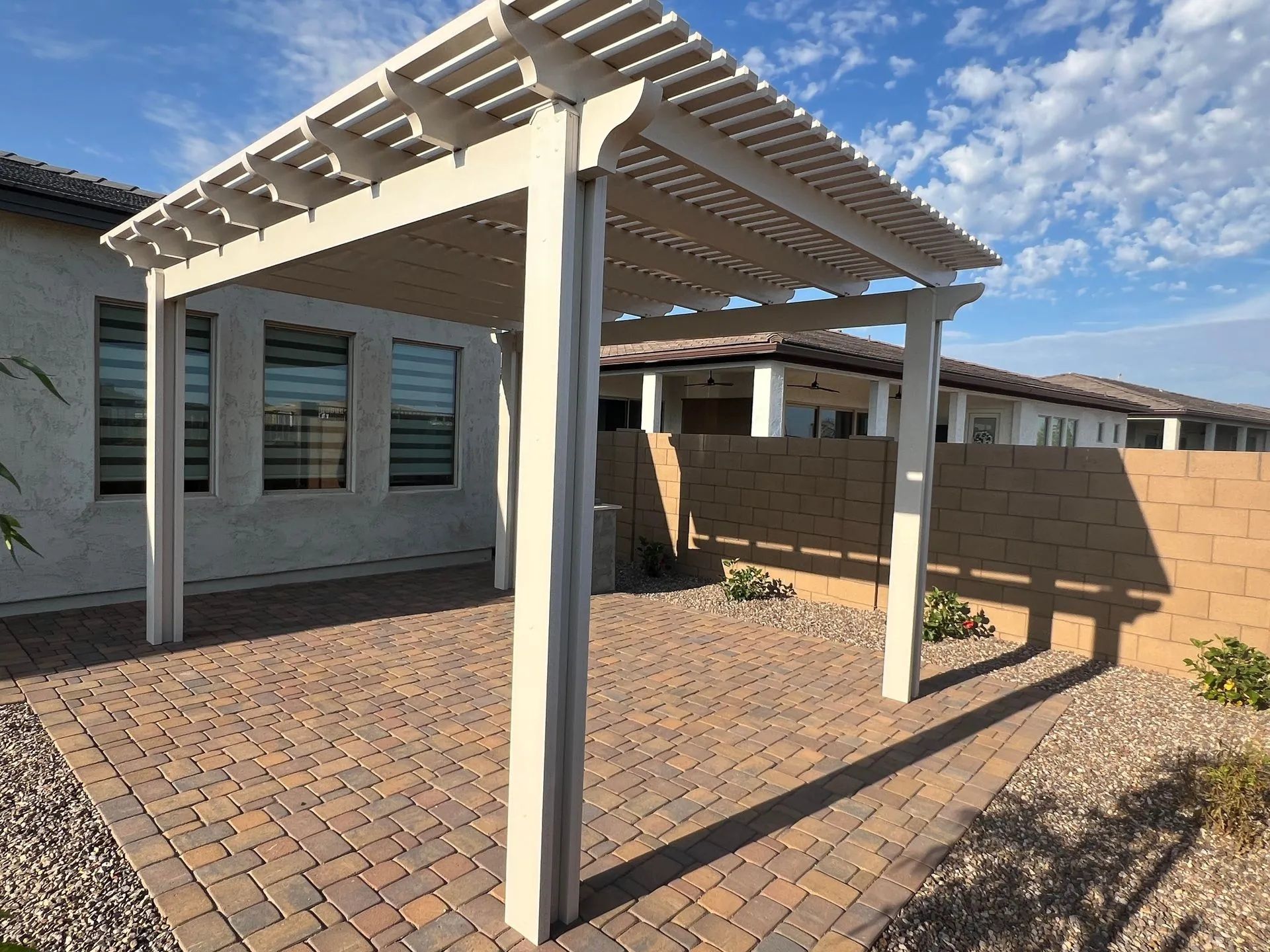 Beige pergola on brick patio; adjacent to a light-colored wall, with a partly cloudy sky.