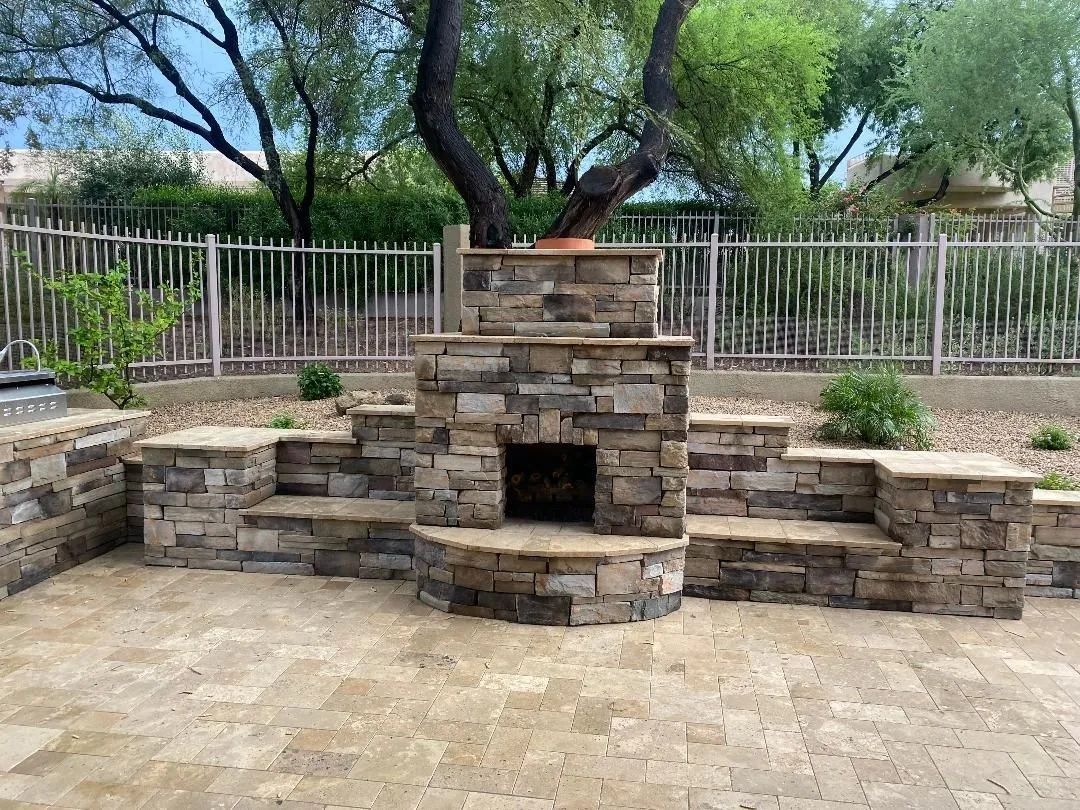 Stone fireplace and seating area in a backyard patio, with trees and a fence in the background.