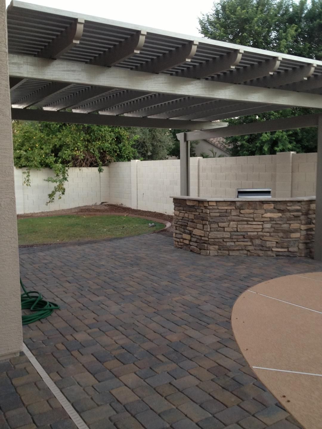 Patio with stone pavers, pergola, built-in grill, and a grassy area against a tall wall.