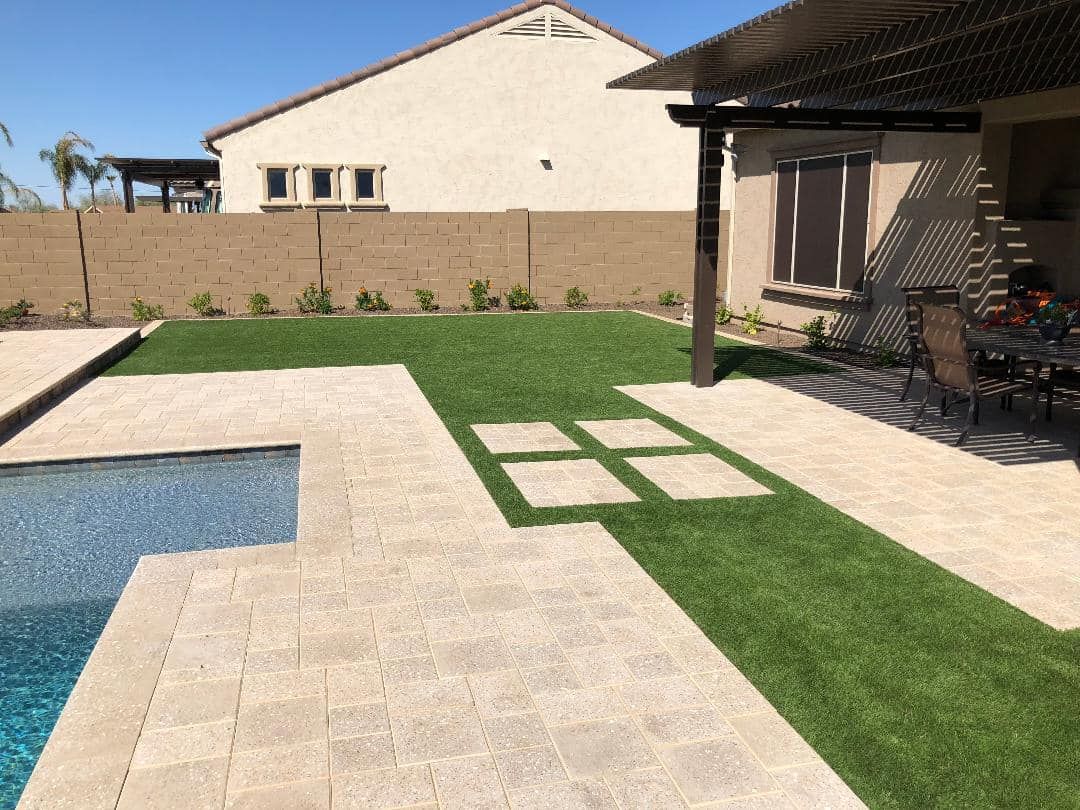 Backyard with pool, paved patio, and artificial turf lawn. Brown fence, beige house in background.