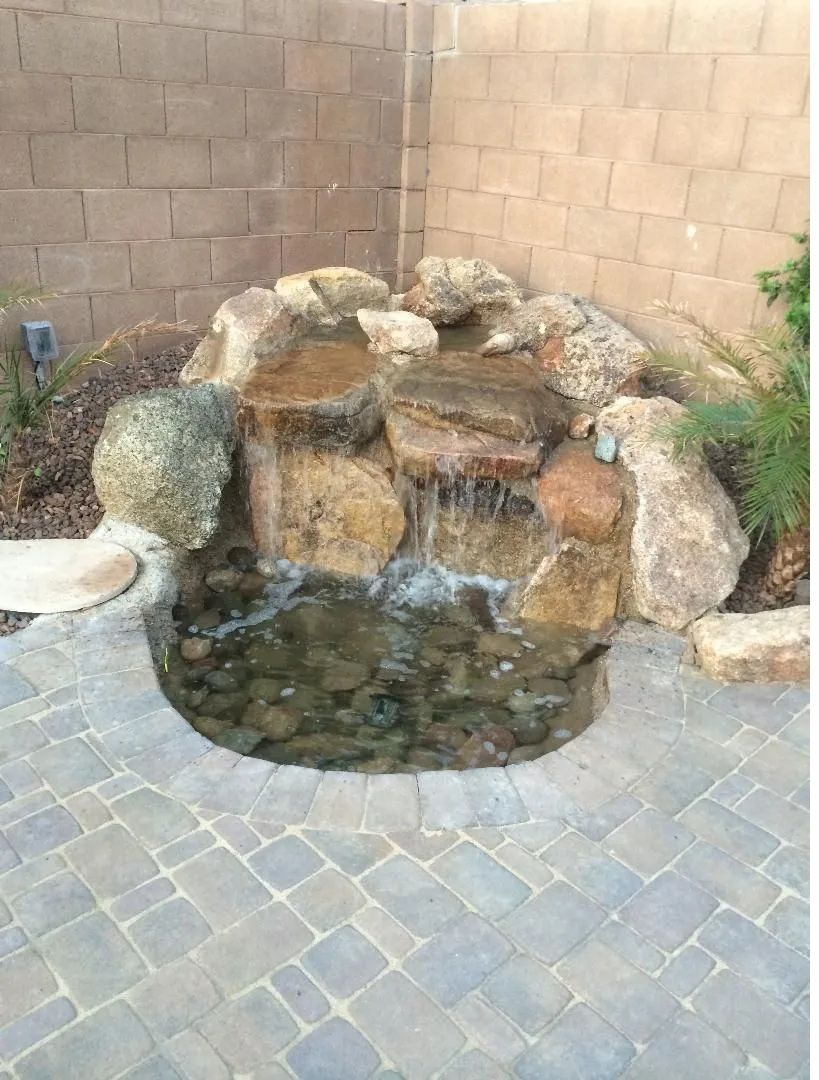 Stone waterfall feature in a backyard, cascading water into a circular pond, surrounded by rock and brick.