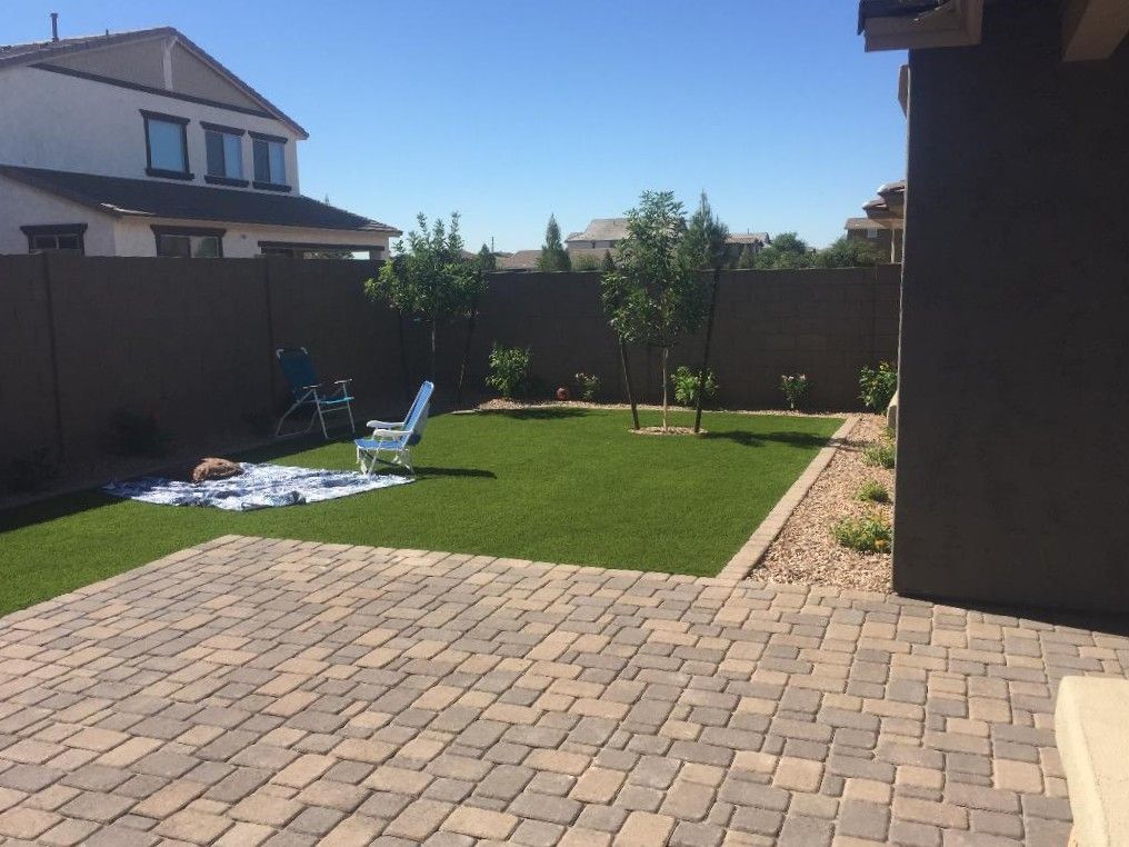 Backyard with paved patio, green lawn, brown fence, and trees under a clear blue sky.