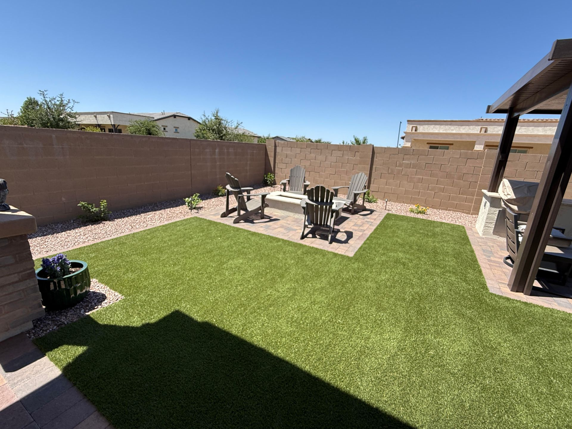 Backyard with green turf, fire pit, chairs, and brick patio. Brown wall and pergola.