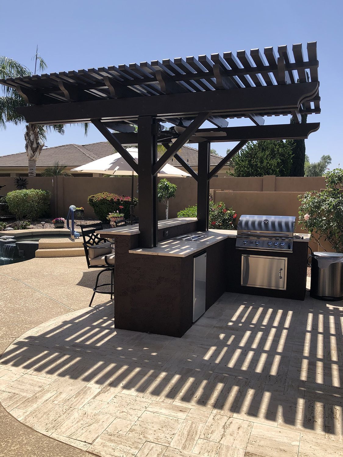 Outdoor kitchen with dark brown pergola, grill, counter, and small refrigerator. Sunny backyard setting.