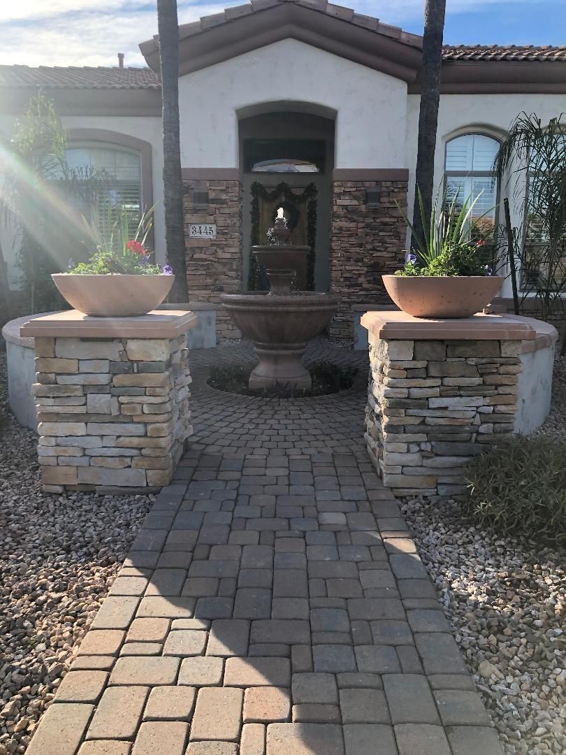 Brick pathway leads to a home with stone pillars holding flower pots. A fountain sits in the center.