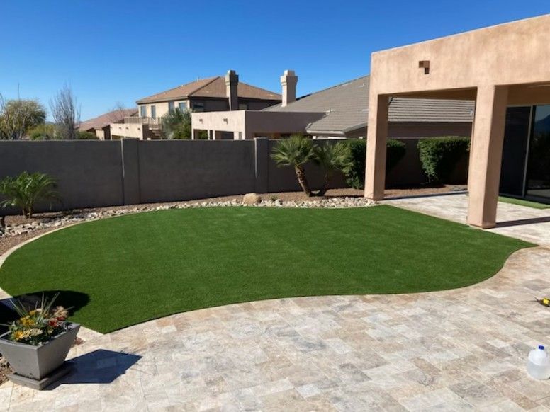 Backyard with green turf, stone patio, and stucco house under a blue sky.