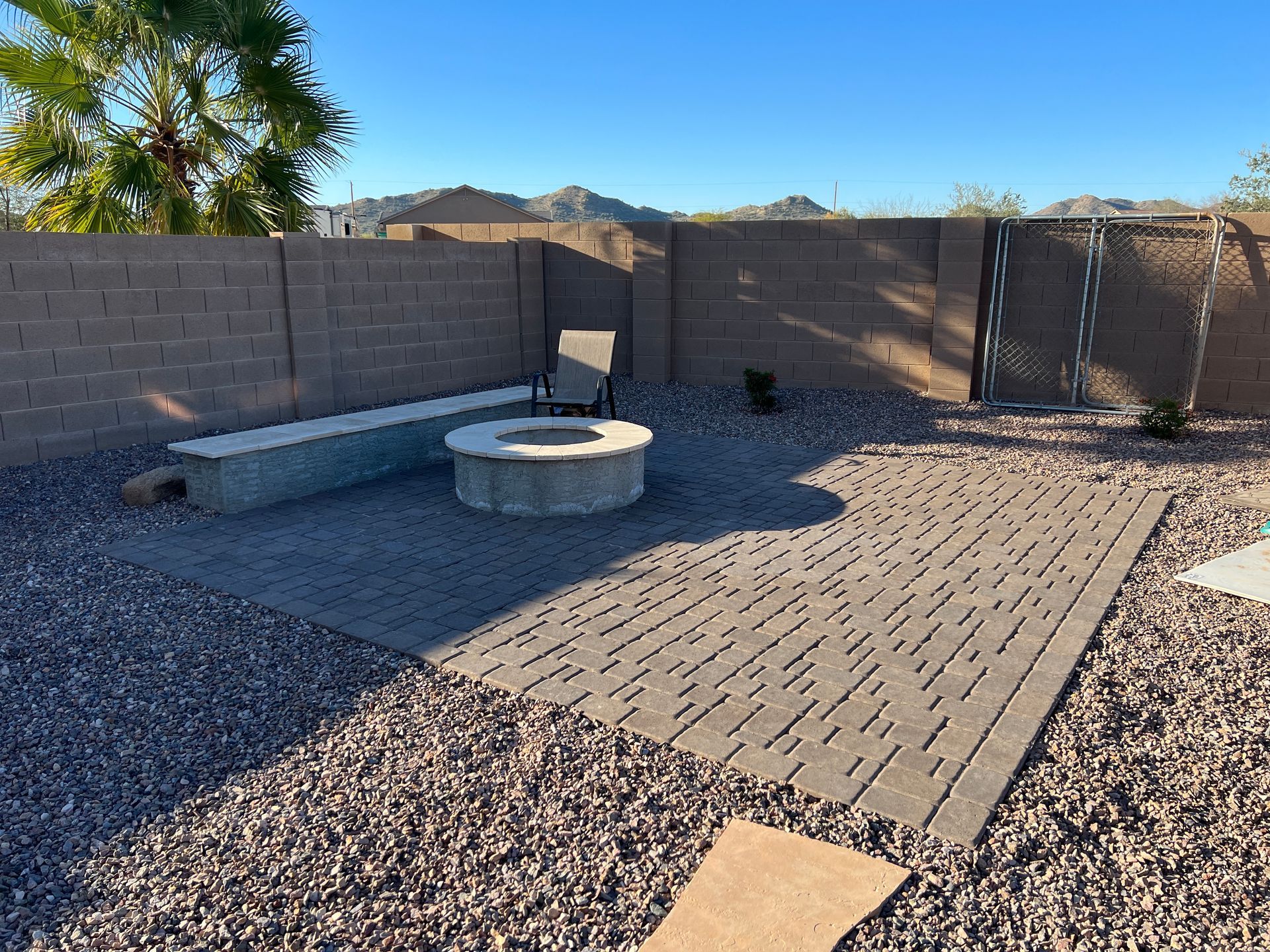 Backyard patio with fire pit, bench, and chair against a tall, brown brick wall.