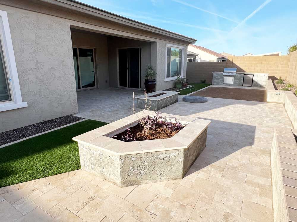 Backyard patio with fire pit, grill, landscaping, beige pavers, and a house.