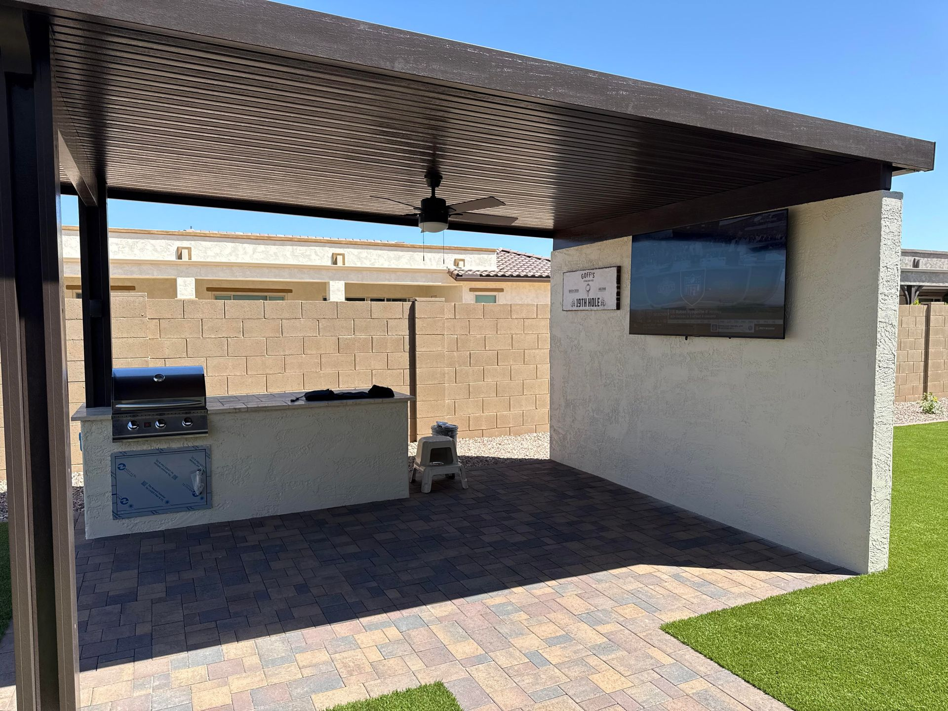 Outdoor kitchen with grill, TV, and seating area under a pergola on a brick patio.